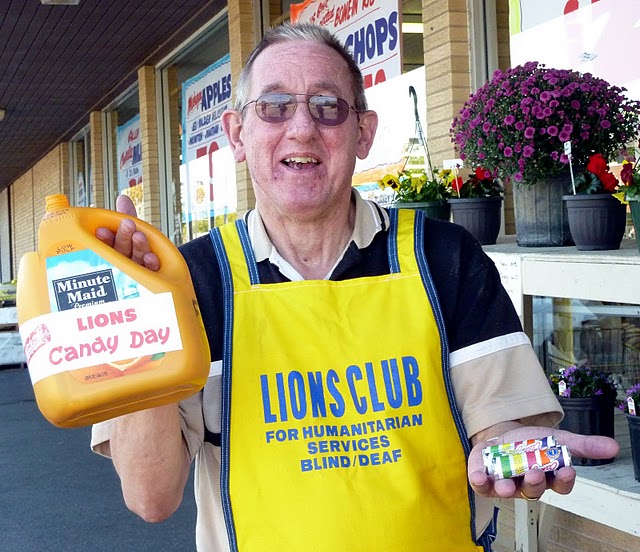 Male Lions Club worker holding a container filled with candy near a storefront, wearing yellow aprons that say: Lions Club Candy Day for the Blind.