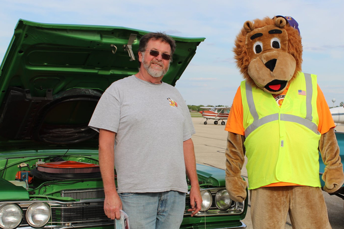 Photo of male and the Lions Club Mascot in front of a green car with an open hood at the Roar-in' Car Cruise.