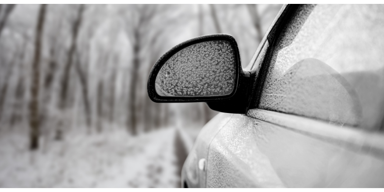 Black and white image of car side mirror covered in snowflakes and frost.