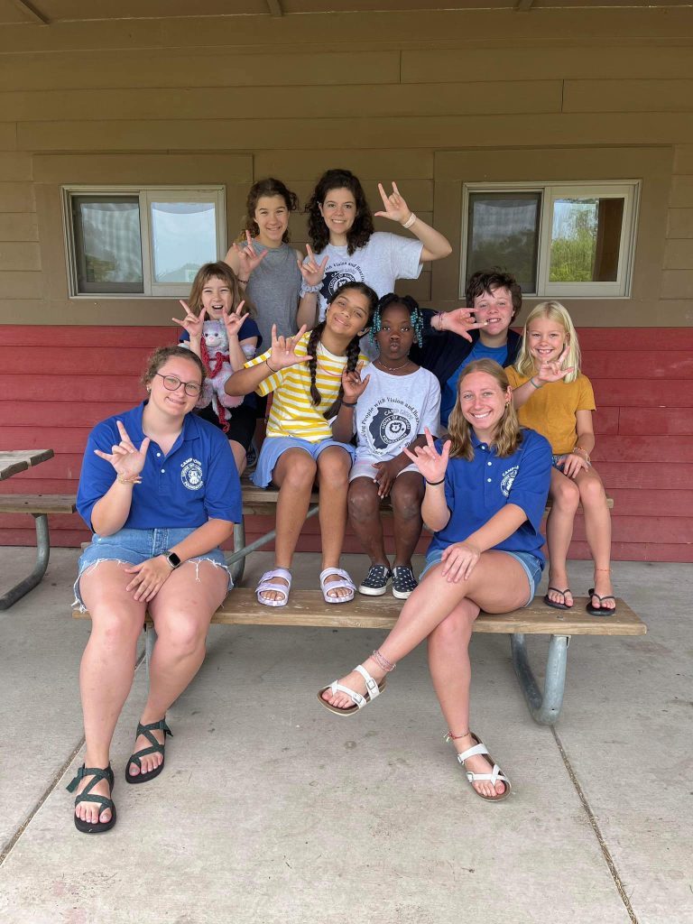 A group of Camp Lions campers and volunteers sit on a picnic bench outdoors and smile at the camera. Some use ASL.