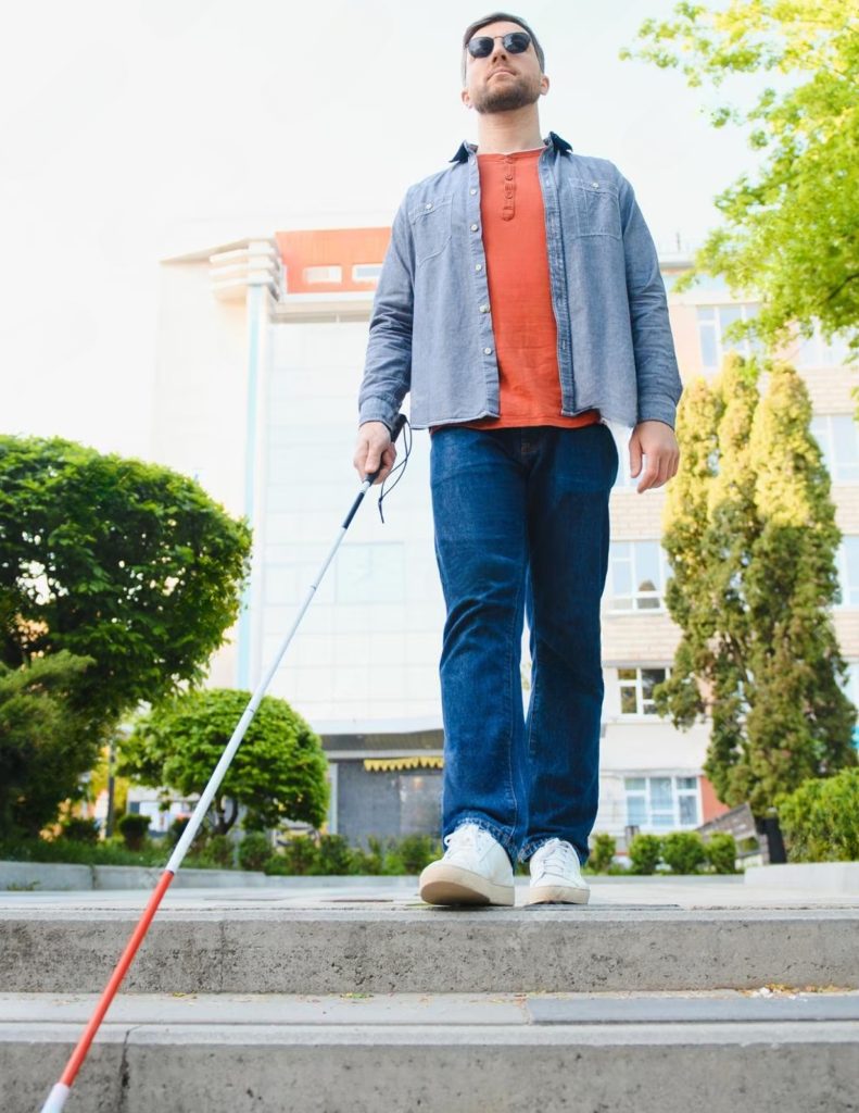 A visually impaired man walking outdoors uses a white cane to navigate a set of steps